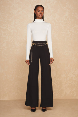 A woman with long braided hair stands against a beige textured background. She is wearing a white long-sleeved top, high-waisted black wide-leg pants, and black pointed-toe heels. The pants are stylishly adorned with the trendy Jane Belt in gold at the waist. She gazes straight ahead.