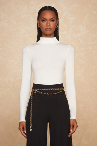 A woman with long braided hair is wearing a white Pearl Giselle Bodysuit featuring a high-neck design, tucked into high-waisted black pants. She accessorizes with a gold chain belt and small hoop earrings. The background is a beige textured wall.