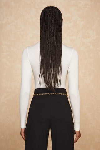 A person with long, neatly braided hair stands facing away from the camera. They are wearing a fitted white long-sleeve top tucked into high-waisted black pants, accessorized with the trendy Jane Belt in gold. The background is a textured beige wall.
