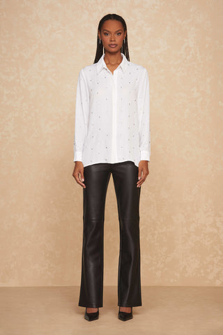 A person with long braids is standing against a beige textured background. They are wearing a white collared Aniya Crystal Shirt from Noli Yoga, adorned with subtle dotted patterns, paired with black form-fitting leather pants and black pointed-toe heels. The shirt features sheer fabric accents, adding an elegant touch to their neutral expression and relaxed posture.