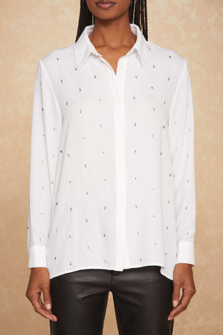 A person wearing the Aniya Top - White, a long-sleeved shirt with small, scattered crystal embellishments and a classic collar. The shirt is paired with black pants against a beige, textured wall. The person's face is not visible.