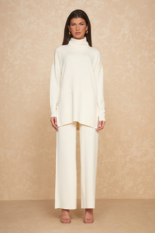 A person stands against a beige textured background, wearing a chic ivory long-sleeved turtleneck sweater known as the Daphne Top - Pearl, paired with matching wide-leg pants and nude heels. Crafted from a silky and breathable soft modal blend, the outfit is accessorized with large gold hoop earrings, perfect for the ultimate on-the-go look.