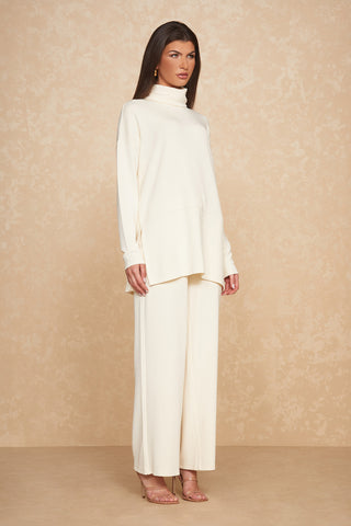 A person with long, dark hair wearing the Daphne Top in Pearl, a light cream-colored, loose-fitting turtleneck sweater made from a soft modal blend, alongside matching wide-leg pants stands against a beige textured backdrop. They are also wearing clear-strap heels and have a neutral expression while looking slightly to the side.