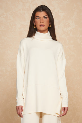 A woman with long dark hair stands against a beige, textured background. She is wearing the Daphne Top in Pearl by Noli Yoga, a cream-colored, long-sleeved turtleneck sweater paired with matching pants that embody luxurious comfort. She has a neutral expression and is accessorized with large gold hoop earrings.