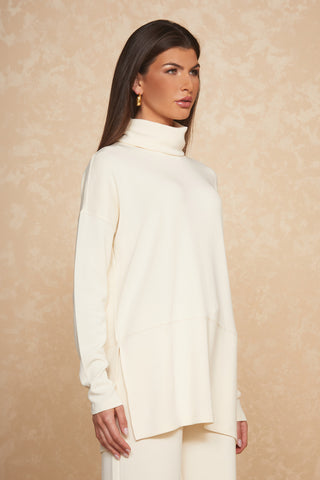 A woman with long, straight brown hair stands against a beige textured background. She is wearing the Daphne Top - Pearl, a cream-colored turtleneck sweater made from a soft modal blend that offers a silky and breathable feel. Matching wide-leg pants complete her ultimate on-the-go look. She has gold hoop earrings and a neutral expression.