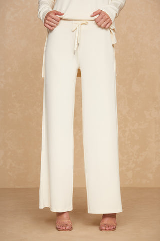 A person wearing the Noli Yoga Daphne Pant in Pearl, featuring a high-waisted, wide-leg design with a drawstring belt, stands against a beige textured background. They are also wearing open-toed beige sandals that epitomize luxurious comfort. The top of their outfit is partially visible.