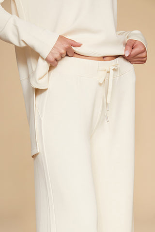 A person wearing the Daphne Pant in Pearl, crafted from a soft modal blend with a drawstring waistband, adjusts the fit. The plain beige background complements this ultimate on-the-go look.