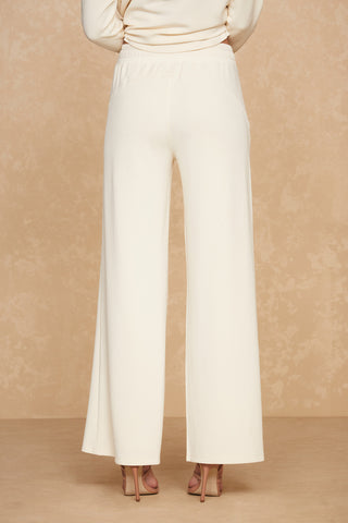 Against a beige background, a person stands with their back to the camera, showcasing the lightweight, wide-leg Daphne Pant in Pearl, made from a soft modal blend. The person is also wearing high-heeled shoes. The upper part of their body is not visible in the frame.