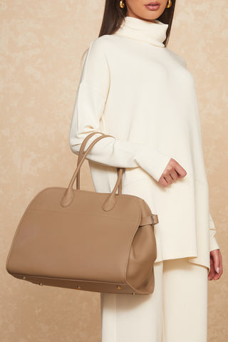 A person is shown from the shoulders down wearing a white turtleneck sweater and white pants, holding a large taupe Margo Bag. The background is a beige textured wall. The outfit and the genuine leather Margo Bag convey a stylish and minimalist fashion sense.