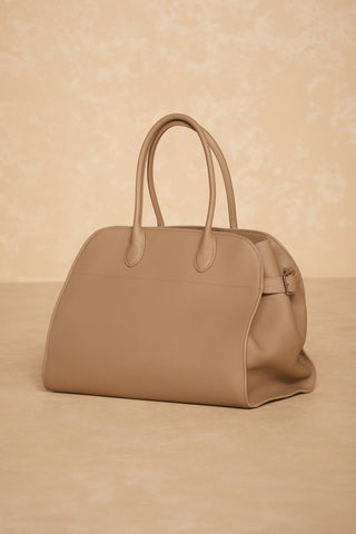 A taupe leather handbag with a classic silhouette and structured design, the Margo Bag from Noli Yoga, is placed against a matching beige background. Crafted from genuine leather, this elegant bag features two sturdy handles and minimalistic stitching for a sleek appearance.