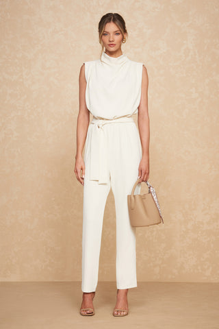 A woman stands against a beige textured background, dressed in a sleeveless white jumpsuit featuring a draped neckline and tied waist. She carries the genuine-leather Athena Bag in taupe and dons beige open-toe heels. With her hair styled in a loose updo, she exudes modern classic elegance as she gazes forward.