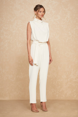 A woman stands against a beige textured backdrop, wearing the flattering Charli Jumpsuit - Pearl with a high neckline and tied waist. She has her hair tied back and is sporting gold hoop earrings and strappy high-heeled sandals.