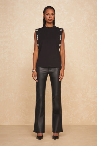 A woman stands against a beige textured background, wearing a sleeveless black Dylan Crystal Top from Noli Yoga with embellished sleeves and decorative buttons on the shoulders and sides. Paired with black leather pants and pointed-toe heels, she has long, braided hair and a composed expression.