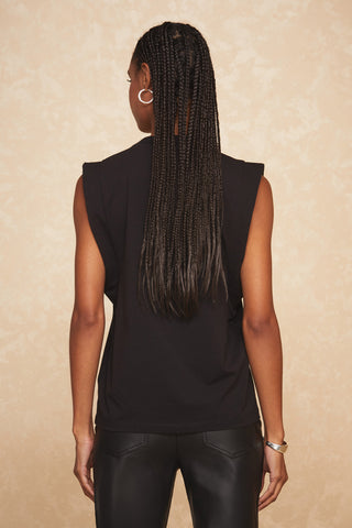 A person with long, braided hair is seen from behind, wearing a sleeveless Dylan Top - Black with embellished sleeves and black leather pants. They have hoop earrings and a bracelet on their left wrist. The background is a beige, textured surface.