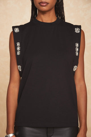 A person wearing a black Dylan Crystal Top by Noli Yoga, featuring embellished sleeves and decorative details along the sides. The background is a light-beige color with a subtle pattern, showing only the individual’s torso, arms, and part of the head.