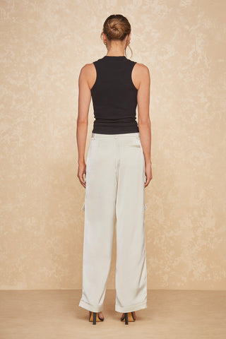 A person with their back to the camera is wearing a black sleeveless top and Milan Satin Cargo Pant in Pearl. They have their hair tied in a bun and stand against a beige, patterned background. The versatile ensemble is completed with black high-heeled shoes.