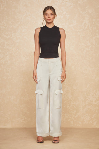 A woman stands against a textured beige wall, wearing the ultra-soft rib Talia Top in black and loose-fitting white cargo pants with multiple pockets. She is also wearing black open-toe heels and has a neutral expression, with her hair styled in a loose updo.