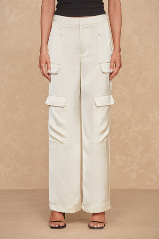 A person is seen from the waist down, wearing the Milan Pant in Pearl, featuring versatile wide legs and two large flap pockets on each thigh. They complement their outfit with open-toe black high-heeled shoes. The background showcases a textured beige wall and floor.