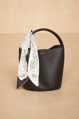 The Athena Bag - Black from Noli Yoga is a textured bucket-style bag featuring a circular handle crafted from genuine leather. A black and white floral twilly scarf is elegantly tied to the handle, creating a stylish modern classic accent against its light beige surface background.