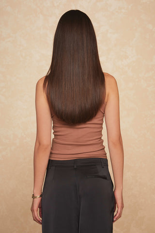 A person with long, straight, dark brown hair stands with their back to the camera. They are wearing the Talia Top - Java, a sleeveless mock neck top that hugs their slim silhouette, paired with dark pants. The background is beige with a subtle textured pattern.