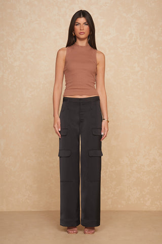 A woman stands against a beige textured wall, wearing the Talia Top in Java—a sleeveless mock neck design—and high-waisted black cargo pants with large pockets. She has long, straight brown hair and is looking directly at the camera. Her slim silhouette is completed with nude heels.