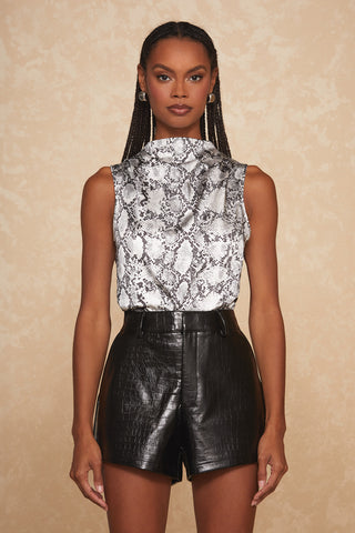 A person with braided hair stands against a beige textured background, wearing the Mara Silk Top - Serpent from Noli Yoga—a sleeveless mock neck featuring a black and white snake print—paired with black, high-waisted, faux leather shorts. Silver earrings complement the chic, fashion-forward look.