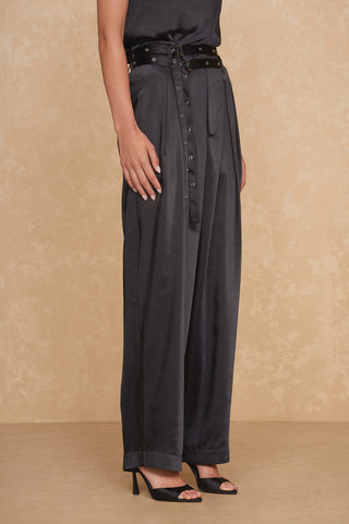 A person is modeling the Kora Pant - Black, high-waisted with a wide-leg cut, paired with a matching black sleeveless top. These satin trousers feature a stylish double belt with multiple grommets and are complemented by black high-heeled sandals. The background is a textured beige wall.