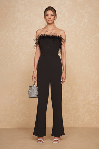 A woman stands against a neutral background, dressed in the Katia Jumpsuit - Black. This strapless jumpsuit features striking feather detailing at the bust. She accessorizes with a small silver handbag in her right hand and silver strappy heels—ideal for party season. Her hair is styled in an elegant updo.