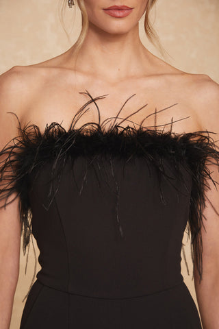 A close-up of a woman wearing the Katia Jumpsuit in black, featuring elegant black feathers along the top hem. Her hair is styled in soft waves, and her face is partially visible with neutral makeup and red lipstick – a true feathered showstopper perfect for party season.