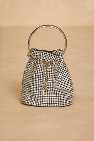 The Celine Bag - Crystal by Noli Yoga is a small handbag embellished with countless glittering crystals, boasting a sleek circular silver handle and a convenient drawstring closure at the top. Set against a solid light tan background, this chic and elegant top handle bucket bag captures attention with its sophisticated design.