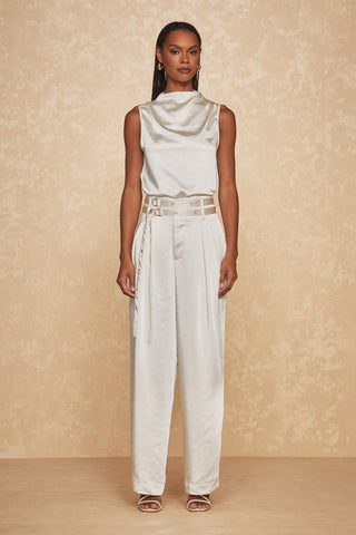 A person stands against a beige textured background, wearing a fashionable outfit that includes the Mara Silk Top - Pearl by Noli Yoga, featuring an effortlessly elegant sleeveless, draped cowl neck. The look is completed with high-waisted, wide-leg trousers, an intricate belt, and matching heeled sandals.