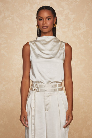 A person stands against a textured, beige background, wearing the effortlessly elegant Mara Silk Top - Pearl by Noli Yoga. The sleeveless top features a draped neckline and is paired with high-waisted trousers that include a matching wide belt adorned with silver eyelets and buckles. Their long, braided hair and hoop earrings complete the sophisticated look.