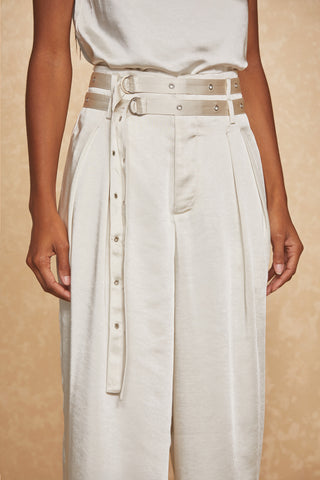 A person wearing the Kora Pant - Pearl, high-waisted textured satin trousers with pleats and a double belt detail, stands against a beige background. The matching belts featuring silver eyelets and buckles add a sophisticated touch to the outfit. The person's arms are relaxed by their sides.