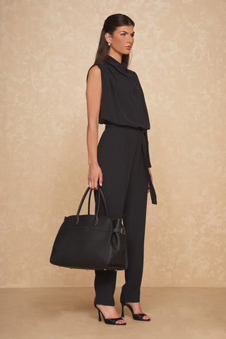 A woman with long, dark hair stands against a beige, textured background. She is wearing a sleeveless, high-neck black jumpsuit and black high heels. She holds the elegant Margo Bag - Black from Noli Yoga in her left hand and gazes off to the side, showcasing a classic silhouette.