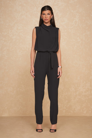 Against a beige, textured background, a woman models the Charli Jumpsuit - Black, a sleeveless slim-fit jumpsuit featuring a cinched waist and mock neck. She styles her long dark hair sleekly back and accessorizes with small hoop earrings, a wristwatch, and black open-toe heels.