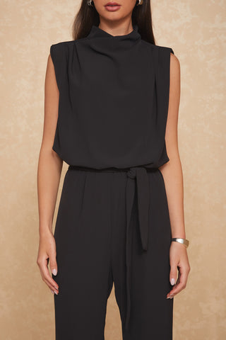 A woman with long dark hair is wearing the Charli Jumpsuit - Black, featuring a sleeveless design, mock neck, and tied waist. She stands against a beige textured background, with a metal bracelet adorning her left wrist.