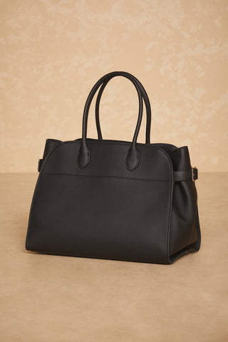 The Margo Bag - Black, crafted from genuine leather, stands out against a beige textured background. Featuring two rounded handles and a structured design with minimal stitching, this sleek handbag offers a sophisticated and stylish look perfect for everyday use.