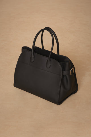 The Margo Bag - Black, a structured black leather handbag with two handles, is placed on a beige surface. Crafted from genuine leather, it boasts a clean, minimalist design with a classic silhouette and smooth texture. A small buckle on one side adds a subtle detail to the accessory, making it perfect for everyday use.