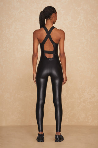 A person with braided hair stands facing backward against a beige textured wall. They are wearing a sleek, black Liquid Vixen Jumpsuit - Black Gloss featuring an open strappy back and black sneakers.