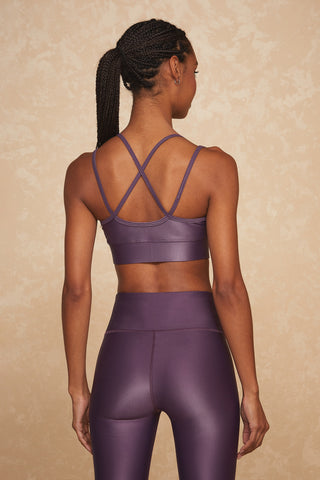 A person with braided hair is wearing a matching high-support Liquid Jolie Bra - Amethyst and leggings, viewed from the back. The sports bra features crossed straps, and the high-waisted leggings are made from liquid fabric with moisture-wicking properties. The background is a neutral, textured tan color.