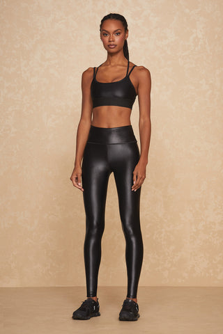 A person stands against a beige textured background wearing a black sports bra and the Liquid Legging - Black Gloss, made from Xtra Life Lycra. They are also sporting black sneakers. Their arms are relaxed by their sides, and their hair is styled in braids.