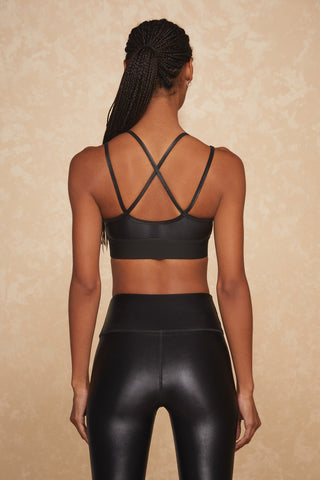 A woman with braided hair stands with her back to the camera against a textured beige background, showcasing the Liquid Jolie Bra - Black Gloss with crisscross straps paired with high-waisted black leggings designed for ultimate comfort through moisture-wicking properties.