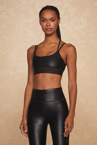 A person stands confidently against a textured beige background, wearing a black Liquid Jolie Bra - Black Gloss and matching high-waisted leggings. With hair styled in braids and a serious expression, they look directly at the camera, showcasing the moisture-wicking design of their outfit.