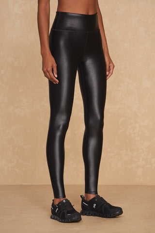 A person is wearing high-waisted Liquid Legging - Black Gloss made from Xtra Life Lycra and black athletic shoes. The photo captures them from the waist down, standing against a beige textured background.