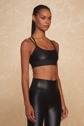 A person is standing against a beige, textured background, wearing a Liquid Jolie Bra - Black Gloss with double shoulder straps and matching high-waisted black workout leggings made from moisture-wicking liquid fabric. They are looking forward with a composed expression and have braided hair.