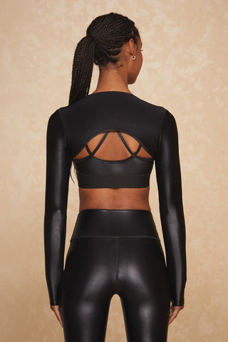 A person with braided hair is standing with their back facing the camera, wearing the Liquid Shrug in Black Gloss—a long-sleeved bolero top with cut-out details—paired with matching black athletic leggings. The background is a neutral, textured surface.