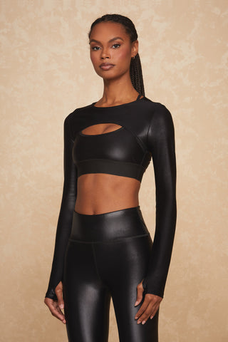 A person with long braided hair is confidently standing against a beige textured background, wearing the Liquid Shrug - Black Gloss by Noli Yoga—a shiny long-sleeved crop top featuring a front cutout and matching high-waisted leggings.