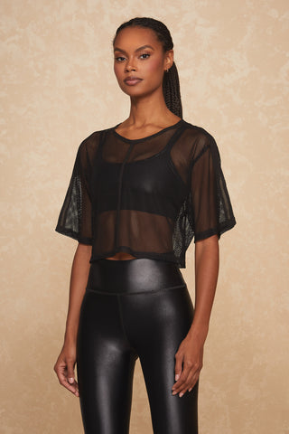 A woman stands against a beige textured background, wearing the Lena Top - Black, a stylish black mesh crop top paired with a matching black sports bra and high-waisted, faux leather leggings. Her long braids cascade down her shoulders as she exudes calm confidence in this chic activewear ensemble.