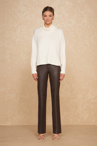 A woman stands against a beige textured background, wearing the Gia Sweater in Pearl, an oversized turtleneck paired with dark brown, high-waisted leather pants. She accessorizes with small hoop earrings and white open-toe heels. Her hair is neatly styled in an updo.