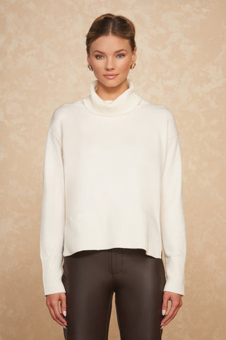 A woman stands against a beige textured background, wearing the Gia Sweater in Pearl, an oversized white turtleneck. She pairs it with black leather pants and has her hair pulled back, accessorizing with gold hoop earrings. She looks directly at the camera with a neutral expression.